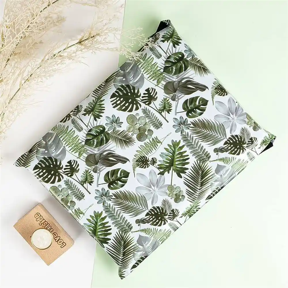 Enveloppe postale écologique au motif de feuilles tropicales vertes, accompagnée d'une petite carte de remerciement en carton, présentée sur un fond vert pastel avec des plantes séchées.